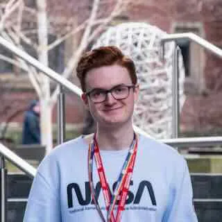 George Wood standing outside in front of some stairs, smiling. He is wearing a pale blue jumper with 'NASA' written across it, and has two lanyards around his neck: one red, and one multicolour.